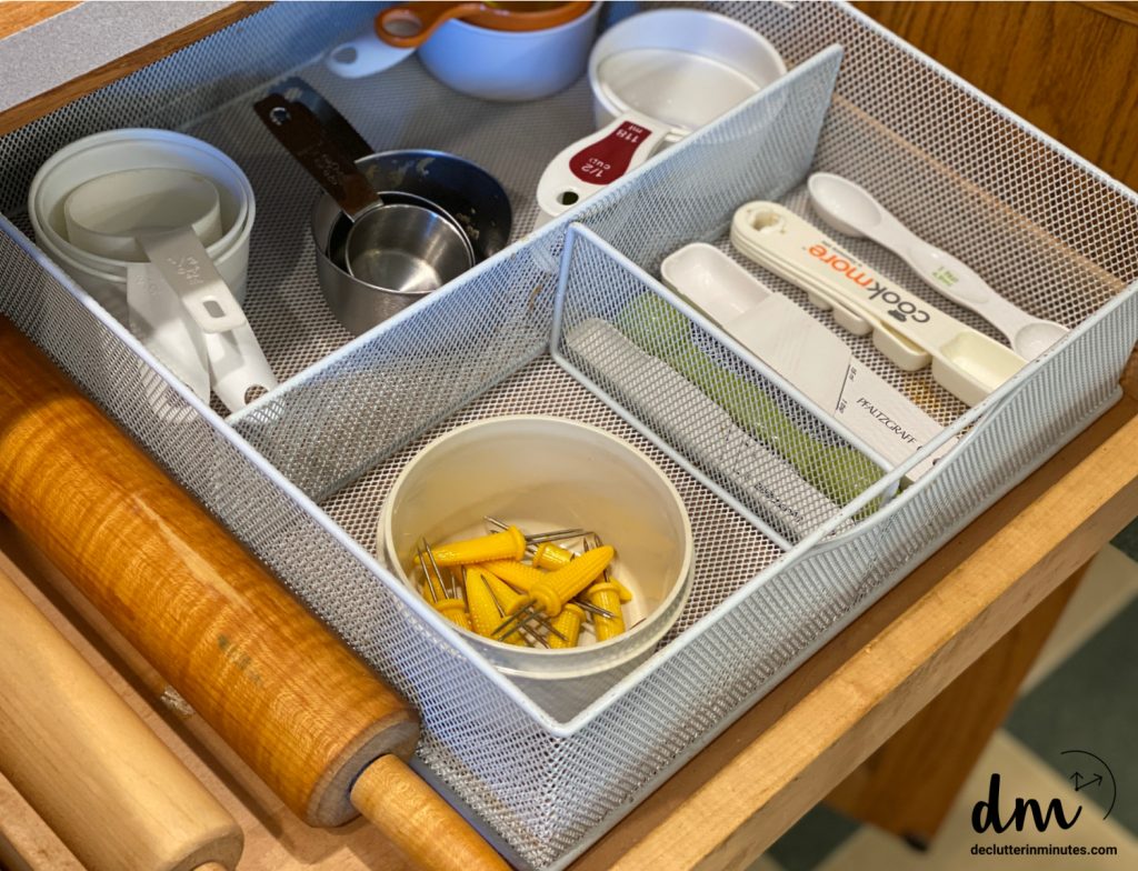 A kitchen drawer with a metal mesh organizer holding measuring cups, spoons, and a rolling pin