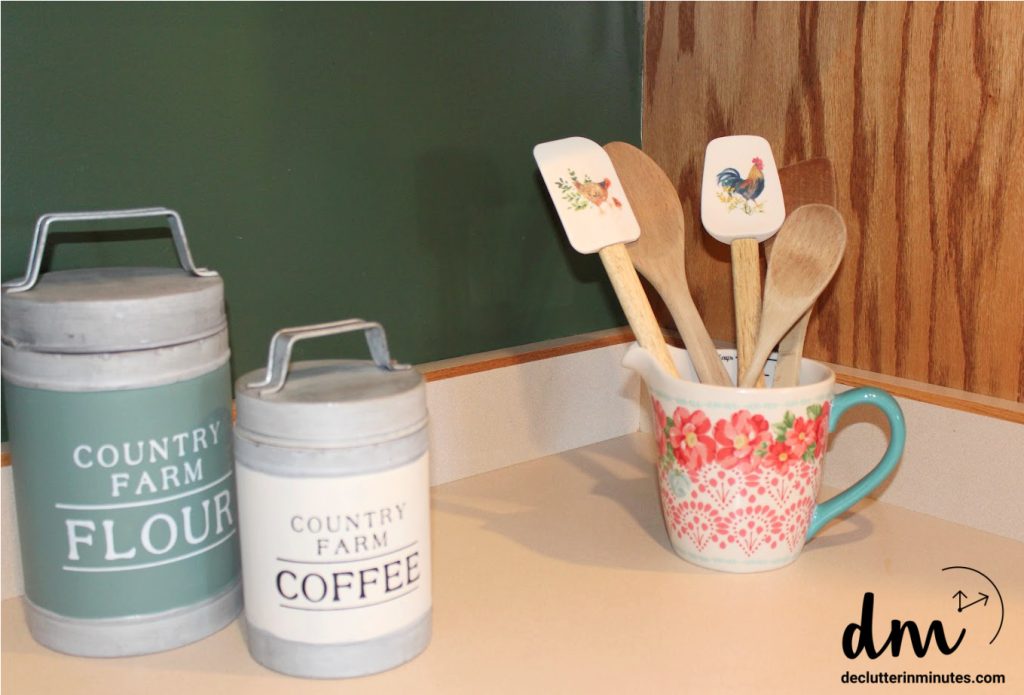 A farmhouse-style kitchen counter with labeled flour and coffee canisters, and a floral mug holding wooden spoons and spatulas with rustic animal prints