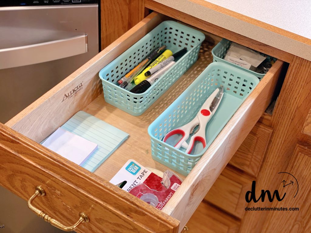An organized kitchen drawer with small plastic baskets holding scissors, pens, markers, tape, and notepads