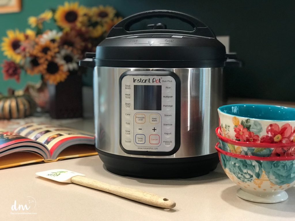 A stainless steel Instant Pot sits on a kitchen counter, surrounded by a cookbook, a wooden spatula, and colorful floral bowls