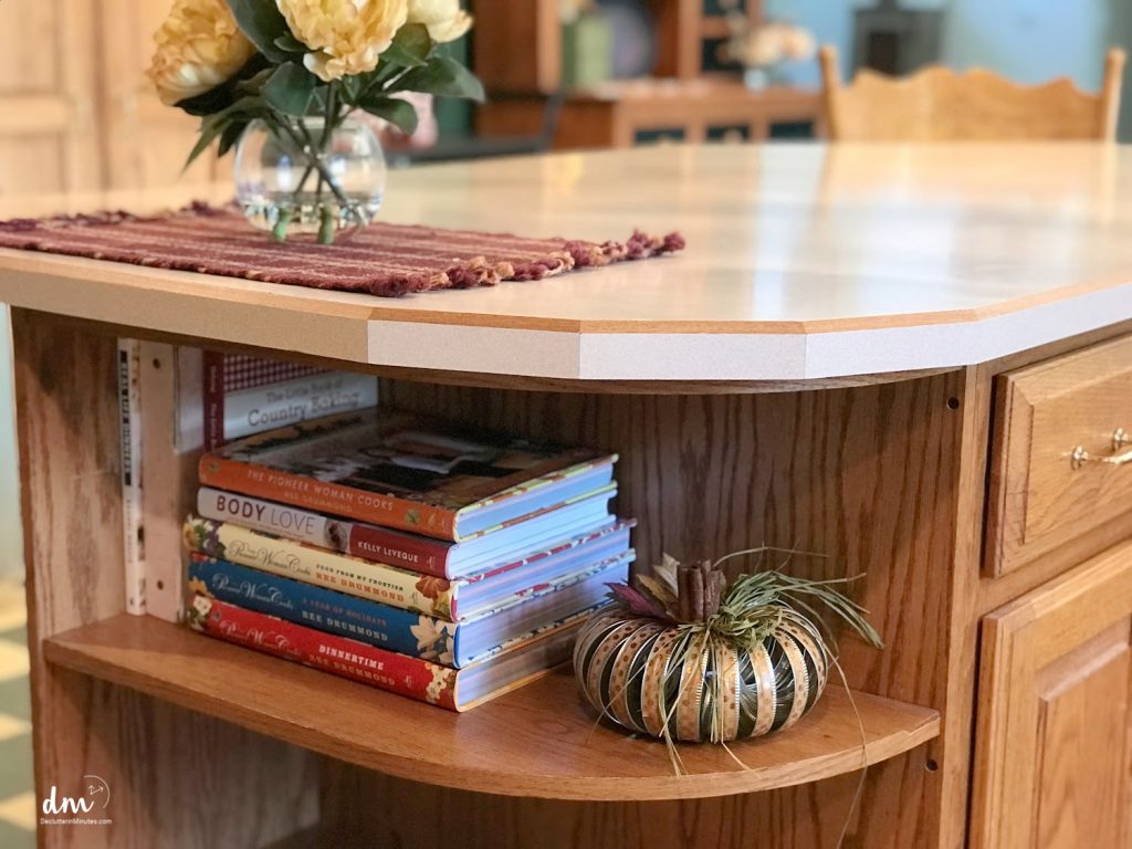 A kitchen island with built-in shelves storing cookbooks, a decorative pumpkin, and a vase with yellow flowers on the countertop