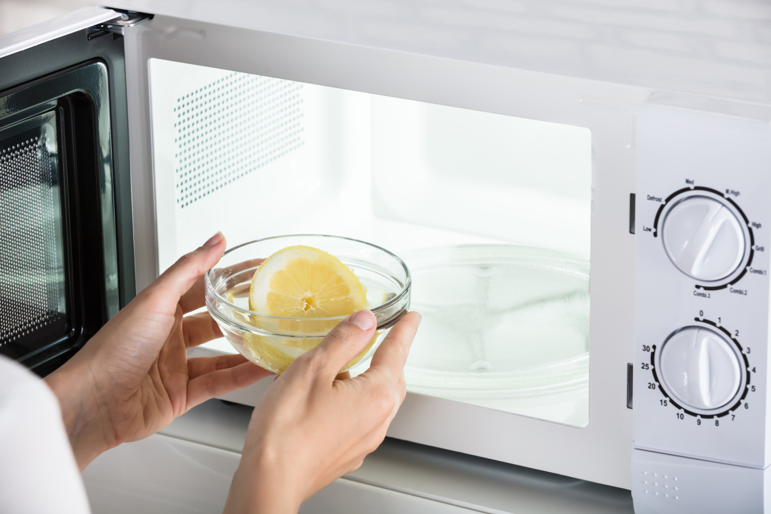 A women placing a glass bowl, with a half of a fruit in it, into a white microwave.