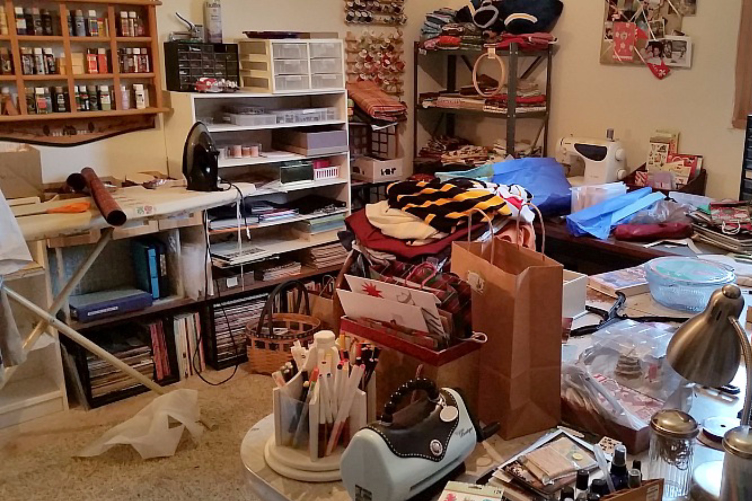 A cluttered craft room filled with supplies, fabric, and tools scattered on shelves, tables, and the floor