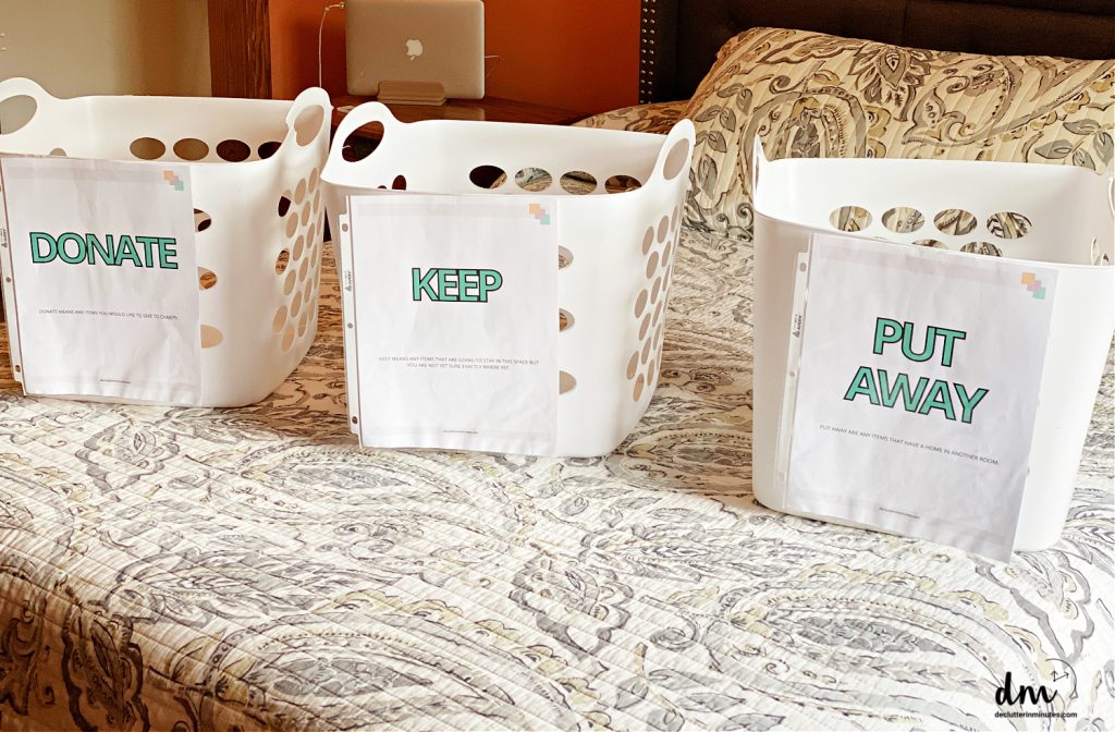 three sorting baskets on a bed with labels on the front that say- Donate, Keep, Put Away 