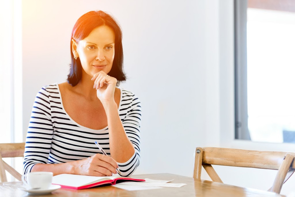 A woman in a striped shirt sitting at a table, writing in a notebook with a cup of coffee nearby
