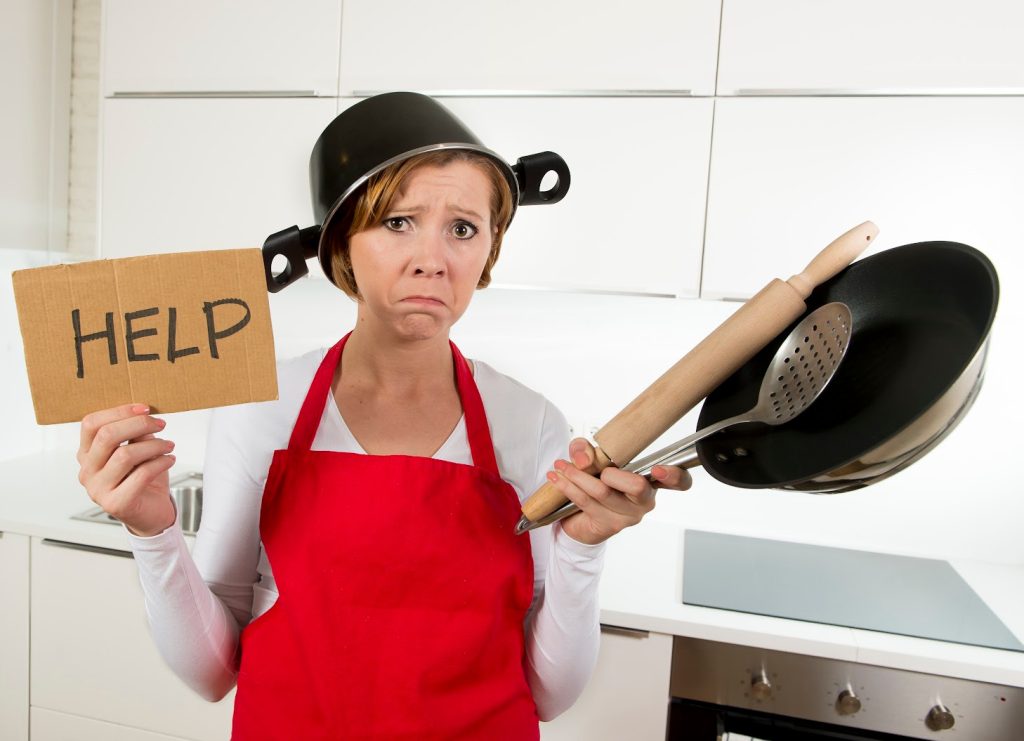 A frustrated woman in a red apron holding a "HELP" sign, wearing a pot on her head, and holding kitchen utensils 