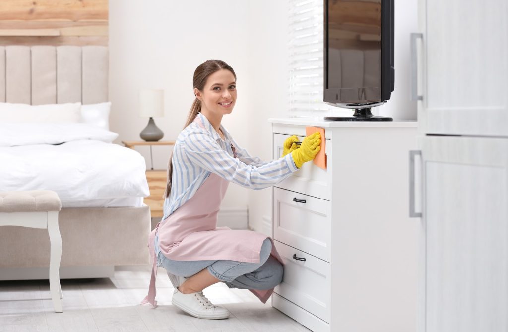 woman wiping dust from the front of a dresser in a bedroom 