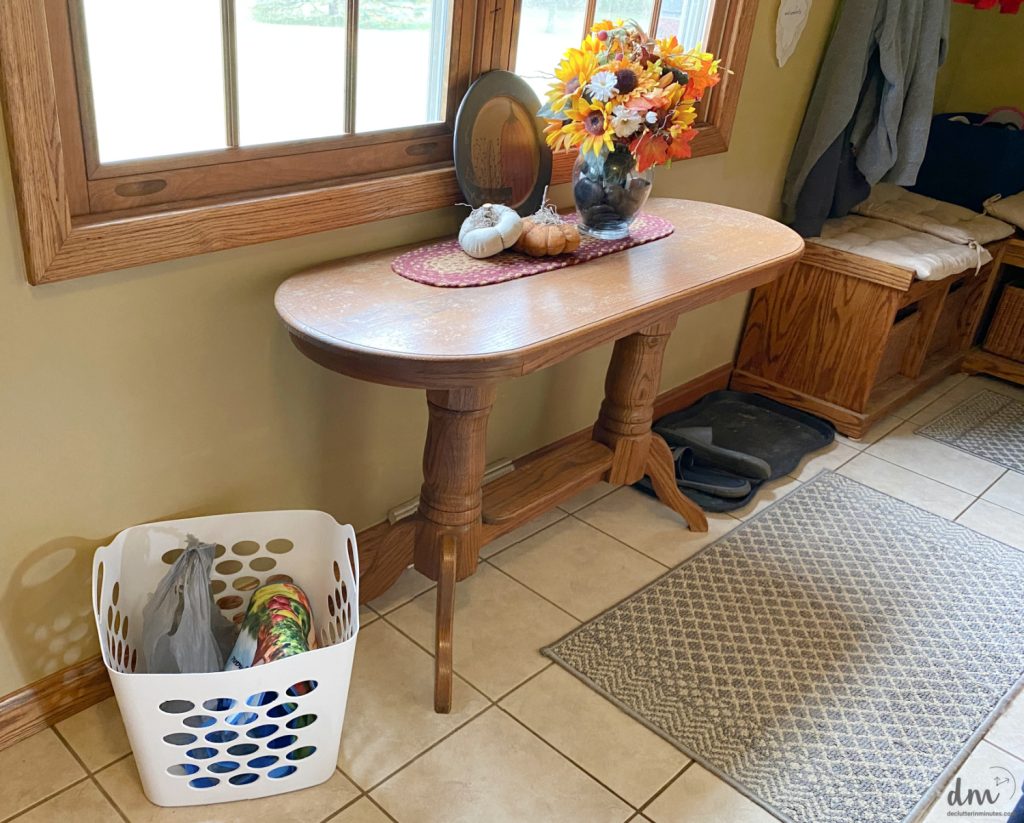 a white basket sitting next to a fall decorated table in a mudroom that is filled with clutter to be put away