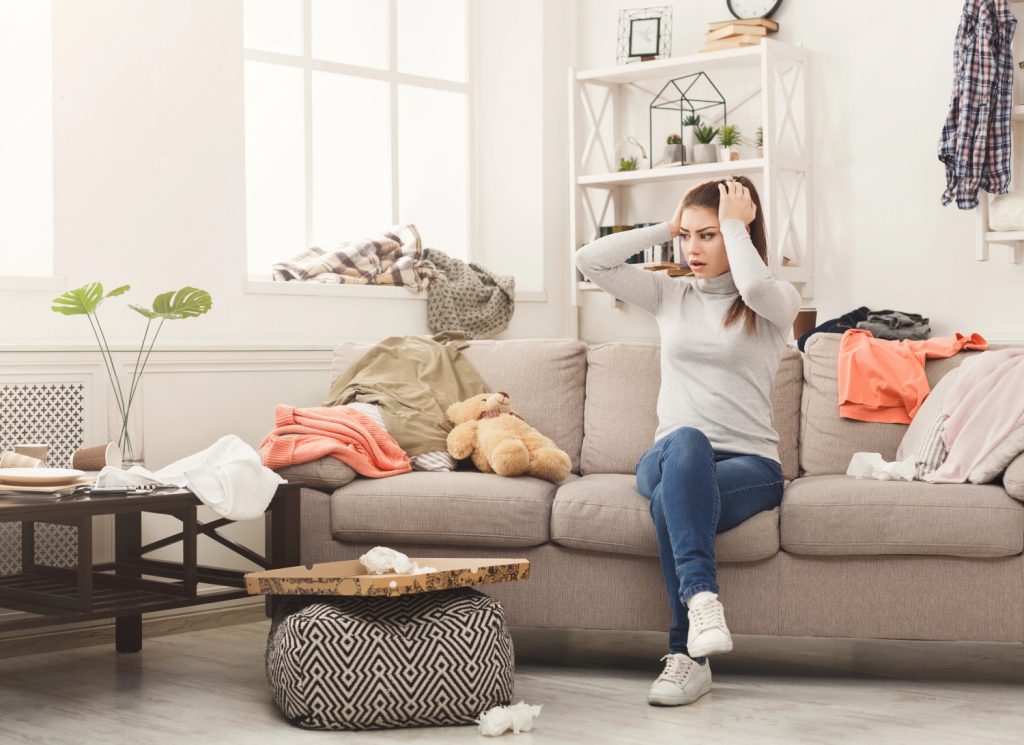 a shocked woman sitting on a couch in a messy and cluttered room 