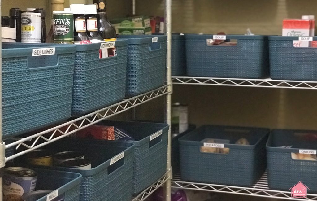 An organized pantry with labeled blue storage bins holding condiments, side dishes, soups, potatoes, and other food items on metal shelves