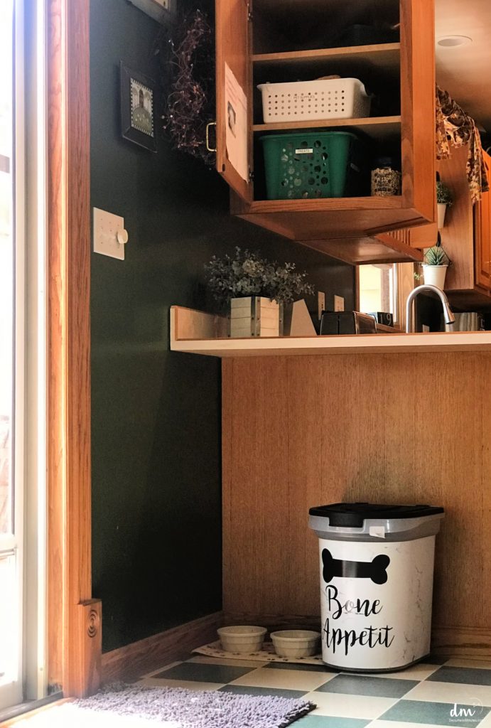 A cozy kitchen corner with an open cabinet containing baskets, a decorative green wall, and a pet feeding area with a bin labeled "Bone Appétit" alongside two bowls