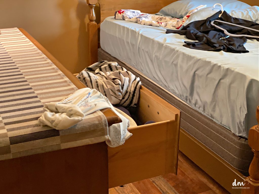 A bedroom with an open storage drawer filled with clothes and scattered clothing on the bed
