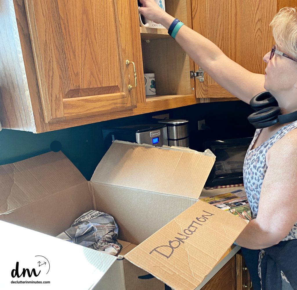 woman in a kitchen decluttering coffee mugs from a cabinet.
