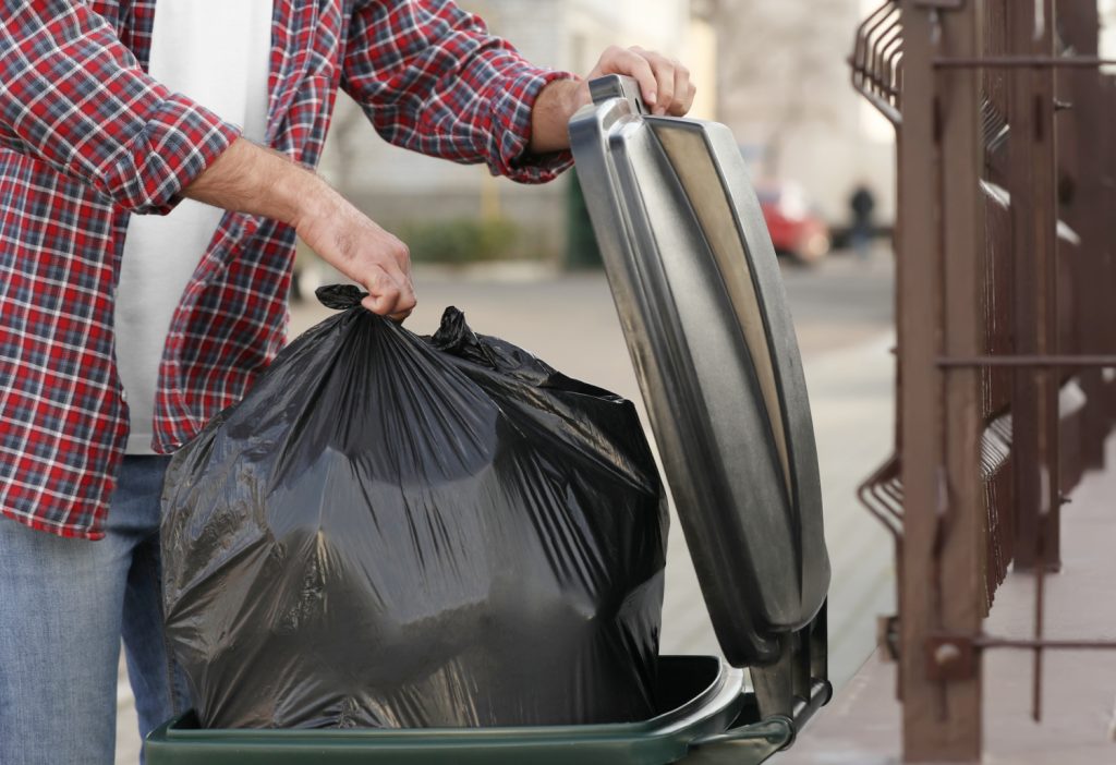 man in red plaid shirt putting black trash bag in a can