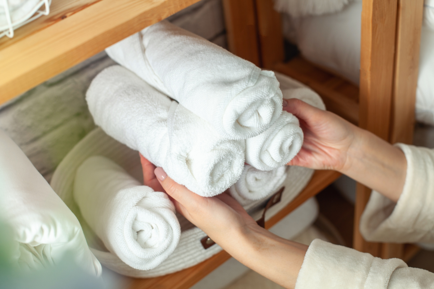 Woman in warm bathrobe is placing basket of rolled cotton bath towels on wooden shelf of linen closet together with bed sheets, blankets, duvet and vase against white brick wall.