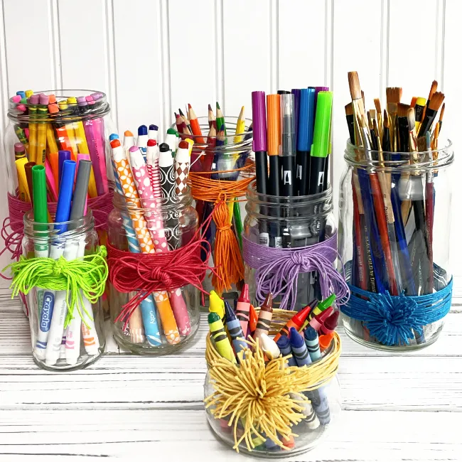 Image that shows: Recycled glass jars tide with string that hold pencils, markers, pencil crayons, markers and paint brushes.