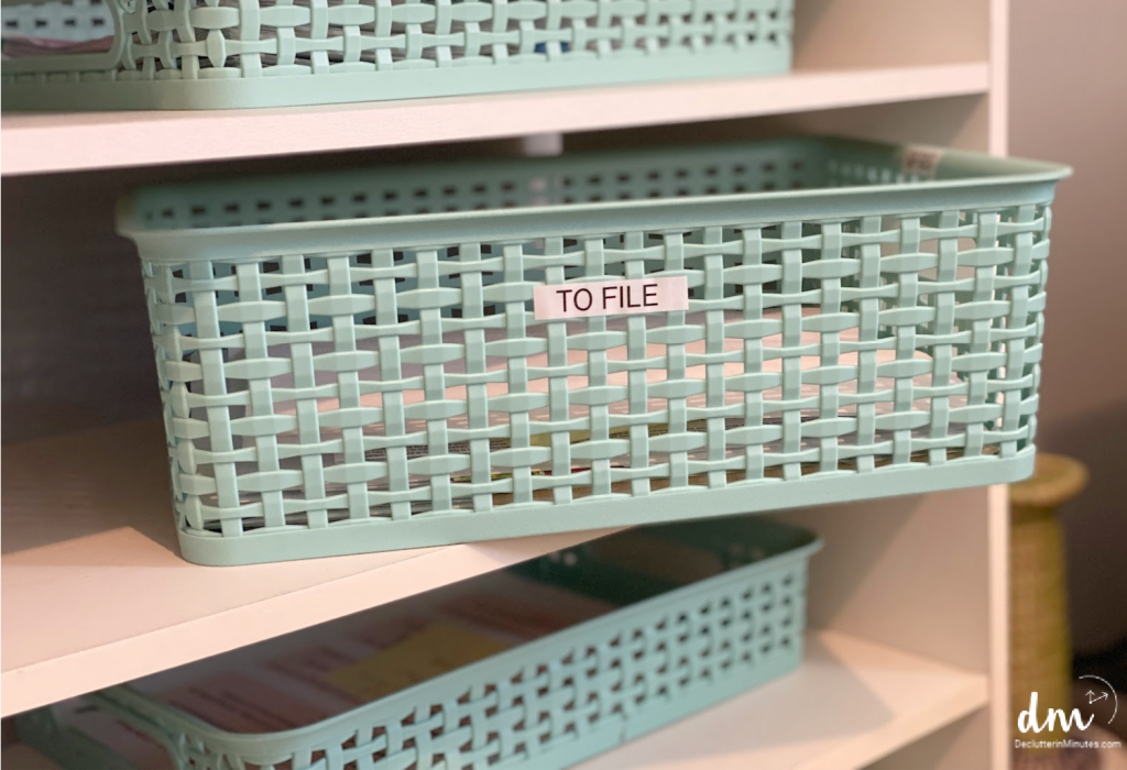 A plastic basket labeled _to file_ on the front sitting on white bookshelf