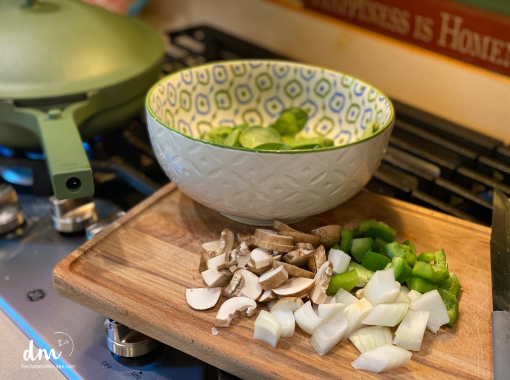 A wooden cutting board on a stovetop holds freshly chopped mushrooms, green bell peppers, and onions, ready for cooking