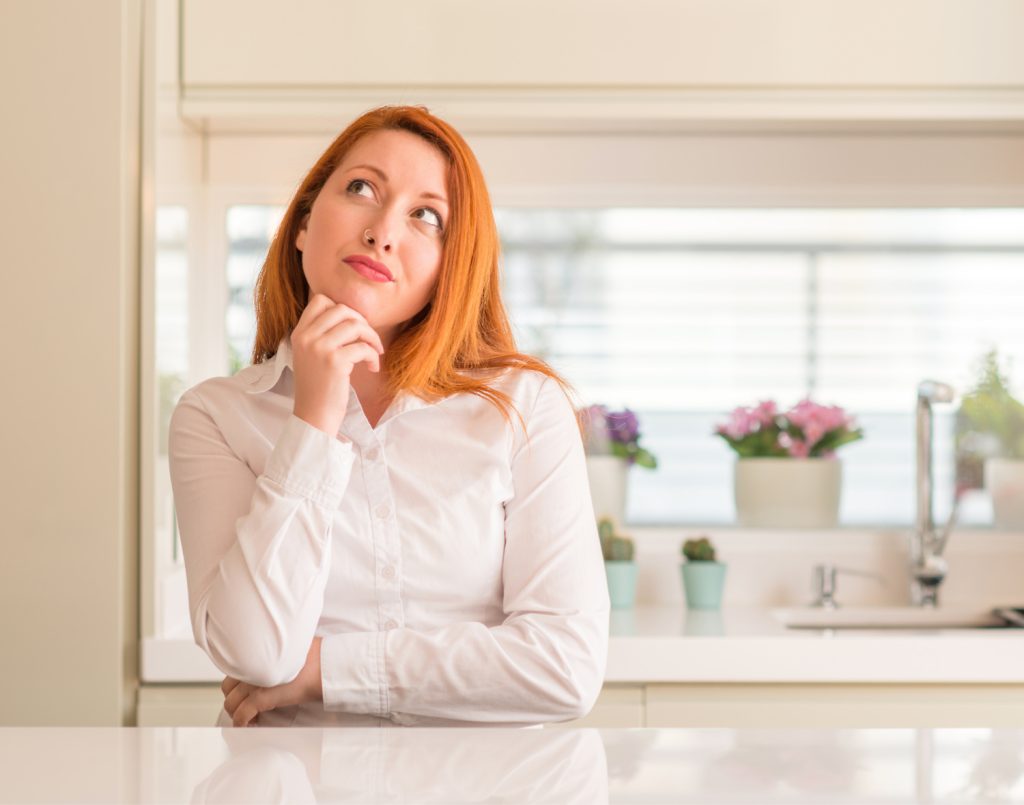 A red-haired woman in a white blouse sits at a kitchen counter, lost in thought
