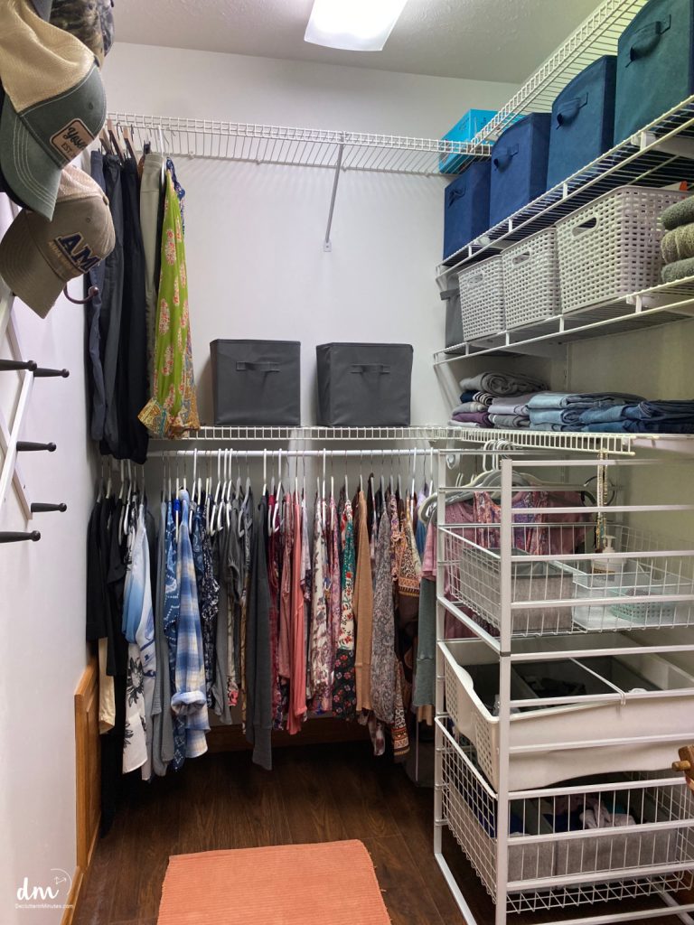 a white closet with wood floors. clothes hanging, hats on a wall, blue and gray baskets on shelves