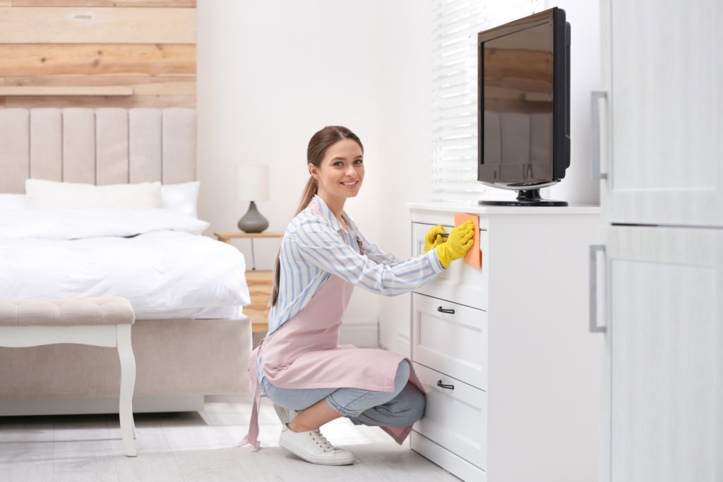 Young chambermaid wiping dust from furniture in hotel room