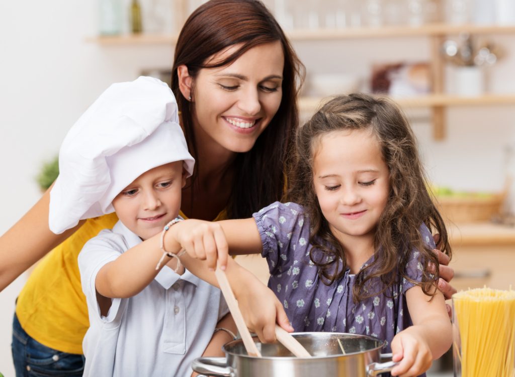 a mom standing behind two young children cooking in a kitchen