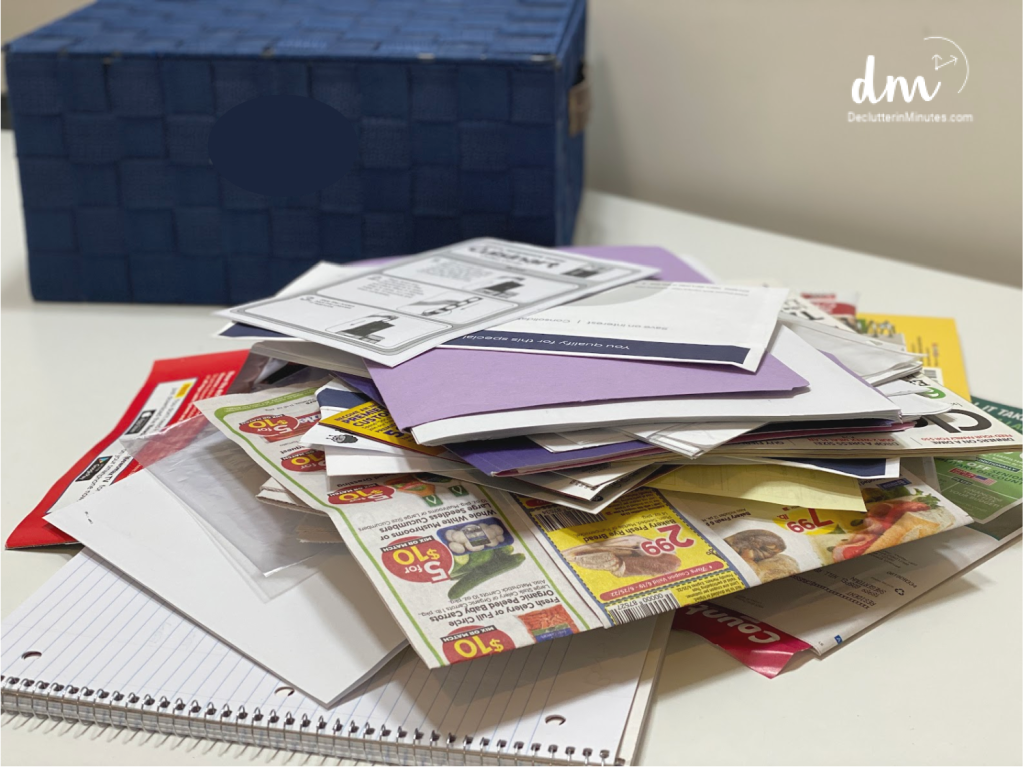A stack of papers, flyers, and notebooks sits on a desk with a blue storage basket in the background