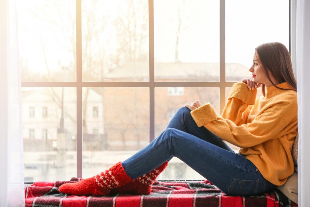 Young woman in winter clothes sitting on window sill at home