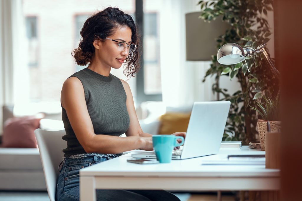 A woman working at a desk in a modern home office, focused on her laptop with a coffee mug nearby
