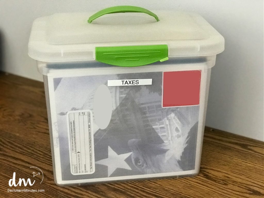 A clear plastic storage box with a green handle, labeled "TAXES" 