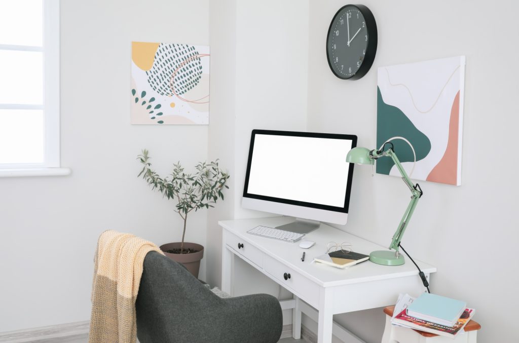 A minimalist home office setup with a white desk, computer, green desk lamp, and modern wall art