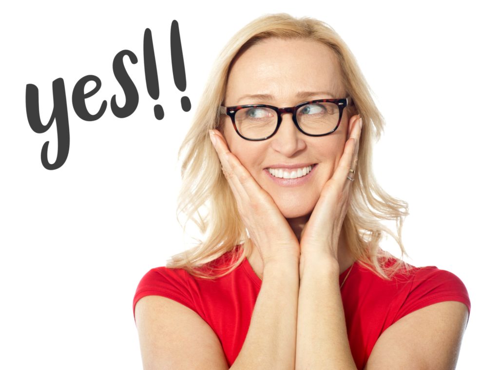 Smiling woman in glasses with hands on her face, excitedly looking to the side with the word 'yes!!' written beside her