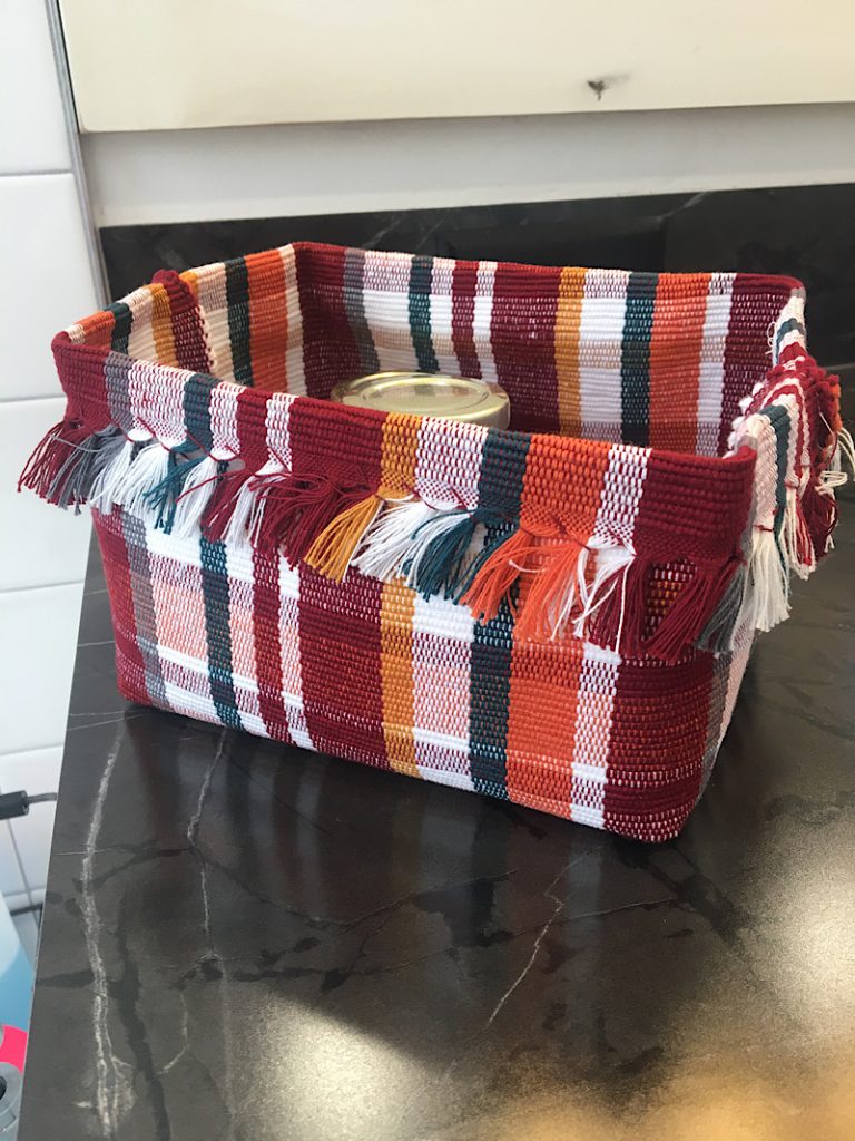 A colorful woven basket with fringe detailing on a countertop