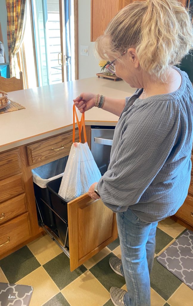 A woman in a blue top removing a tied garbage bag from a pull-out trash bin in a wooden kitchen cabinet