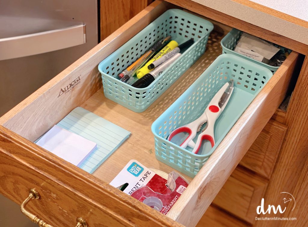 An organized kitchen drawer with light green baskets holding pens, markers, scissors, tape, and notepads