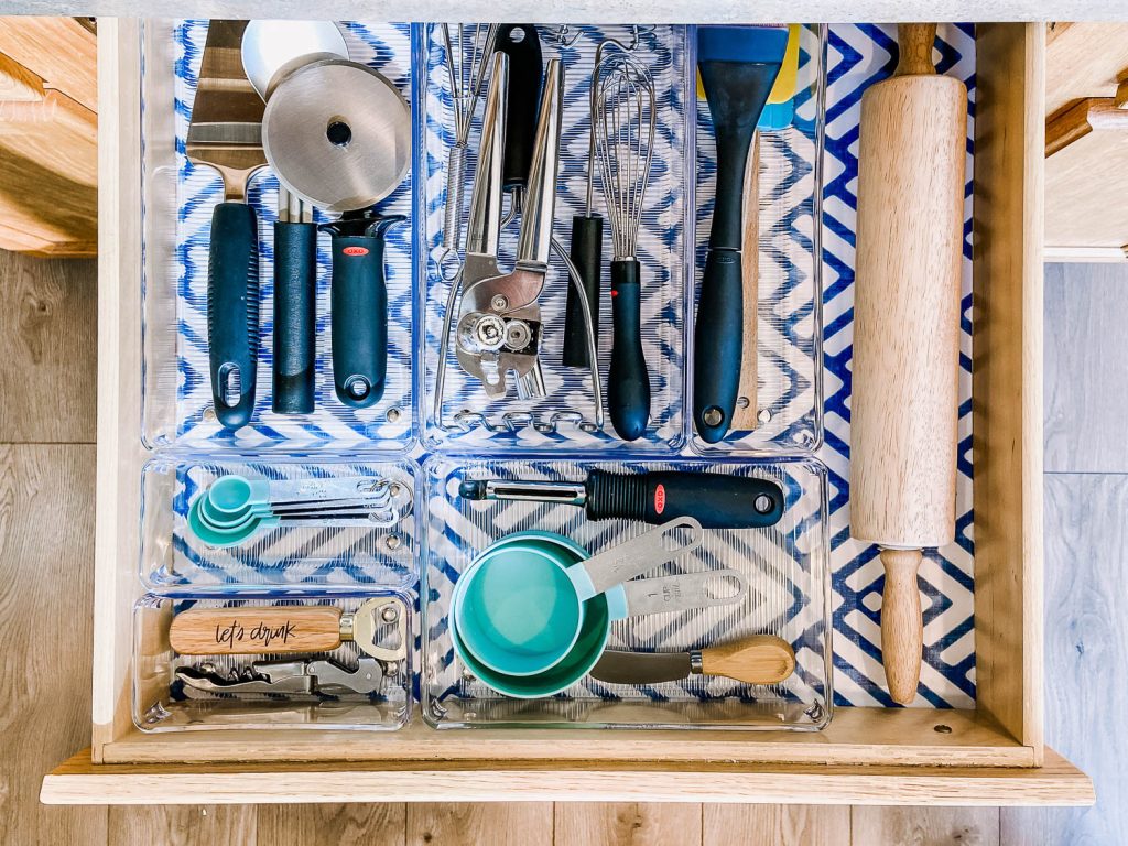 A kitchen drawer with clear dividers holding utensils, measuring tools, and a rolling pin