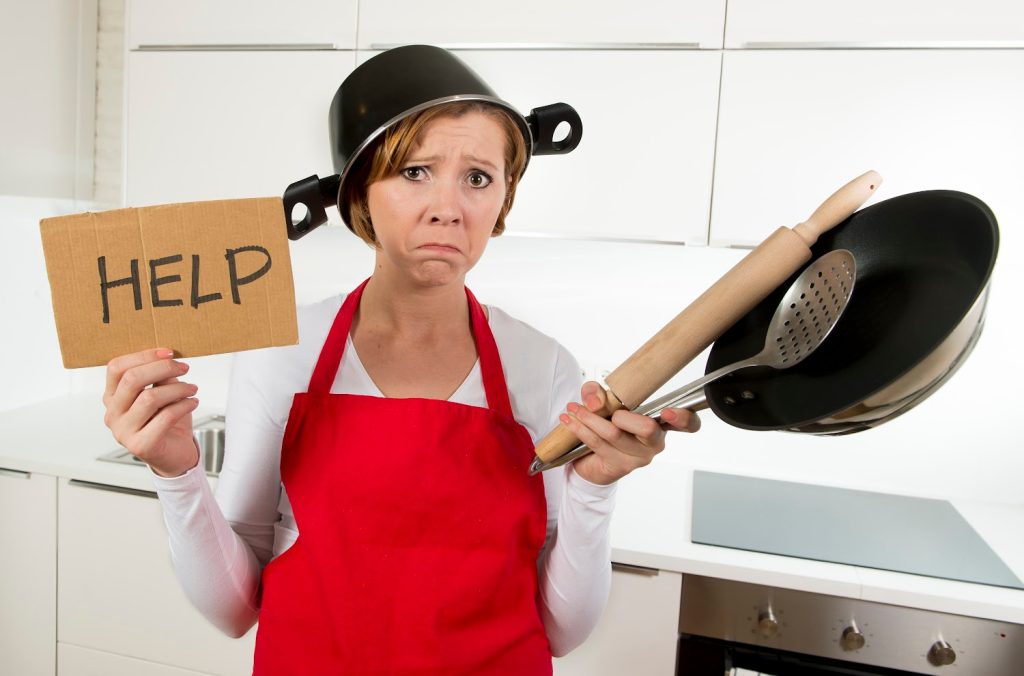 A frustrated woman wearing a red apron holds a "HELP" sign, kitchen utensils, and a pot on her head