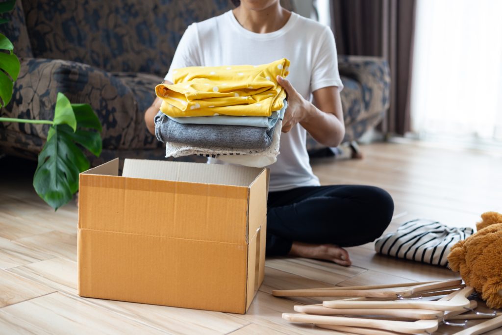 a woman putting clothes in a box for donation hangers on the floor next to her
