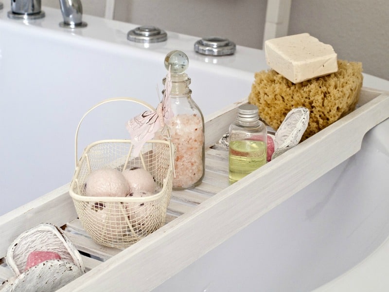 An image that shows a wood tray across the tub holding soap, bath salt, and loofah. 