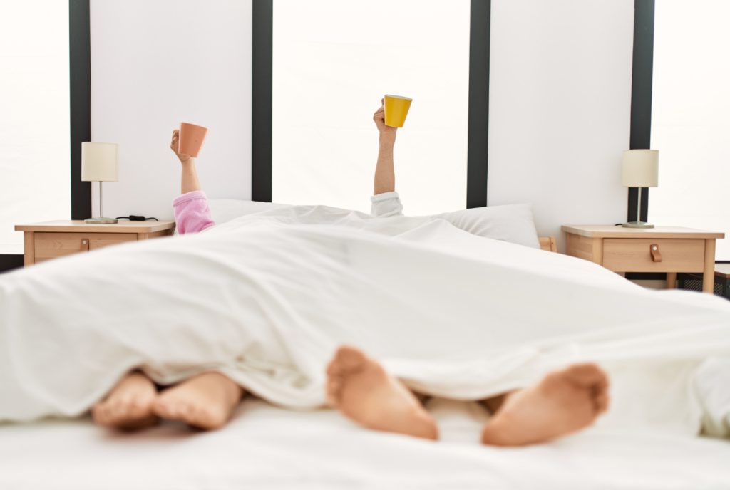 a couple lying on the bed with mug raised up at bedroom.