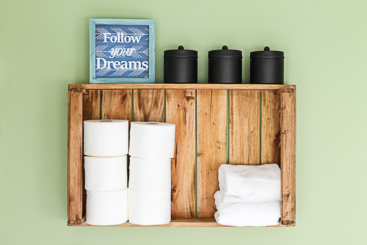 Image that shows a crate over the toilet that holds hand towels, toilet paper, a sign and containers with lids. 