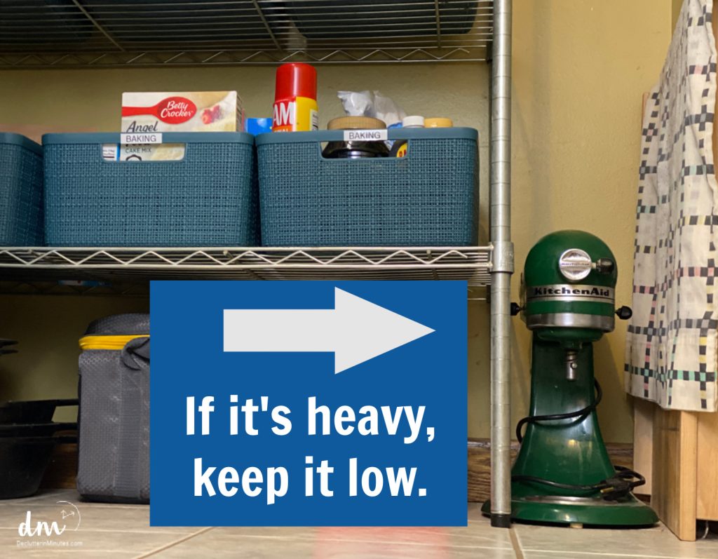 A metal storage shelf with blue baskets holding baking supplies, a green KitchenAid mixer on the floor, and a sign that says, "If it's heavy, keep it low"