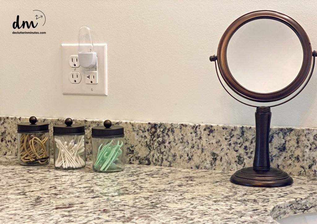 Bathroom counter with glass jars holding hair ties, cotton swabs, and floss picks, next to a small magnifying mirror