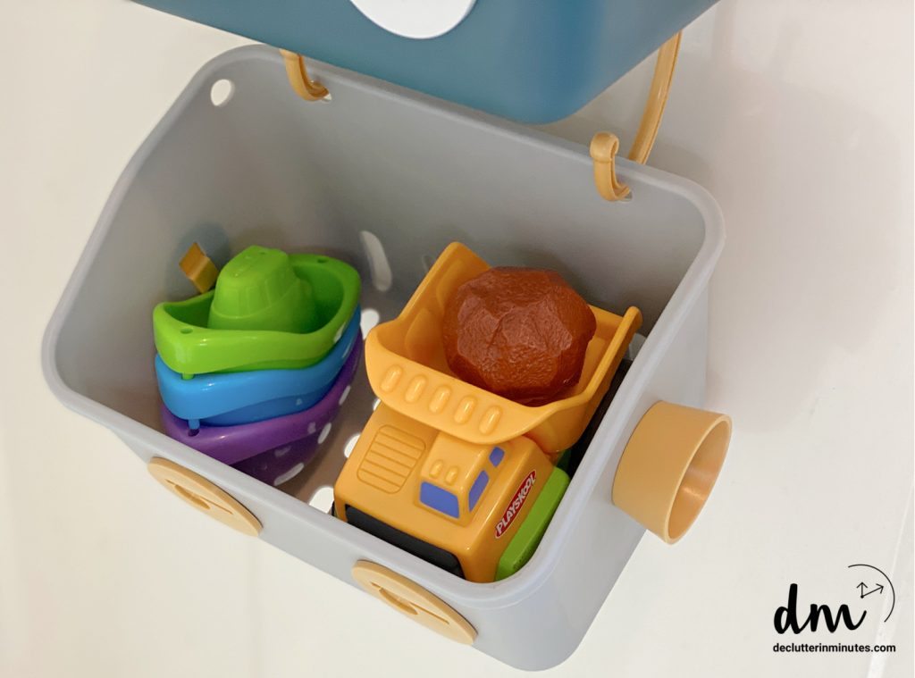 Plastic storage basket holding colorful bath toys, including stackable boats and a toy dump truck
