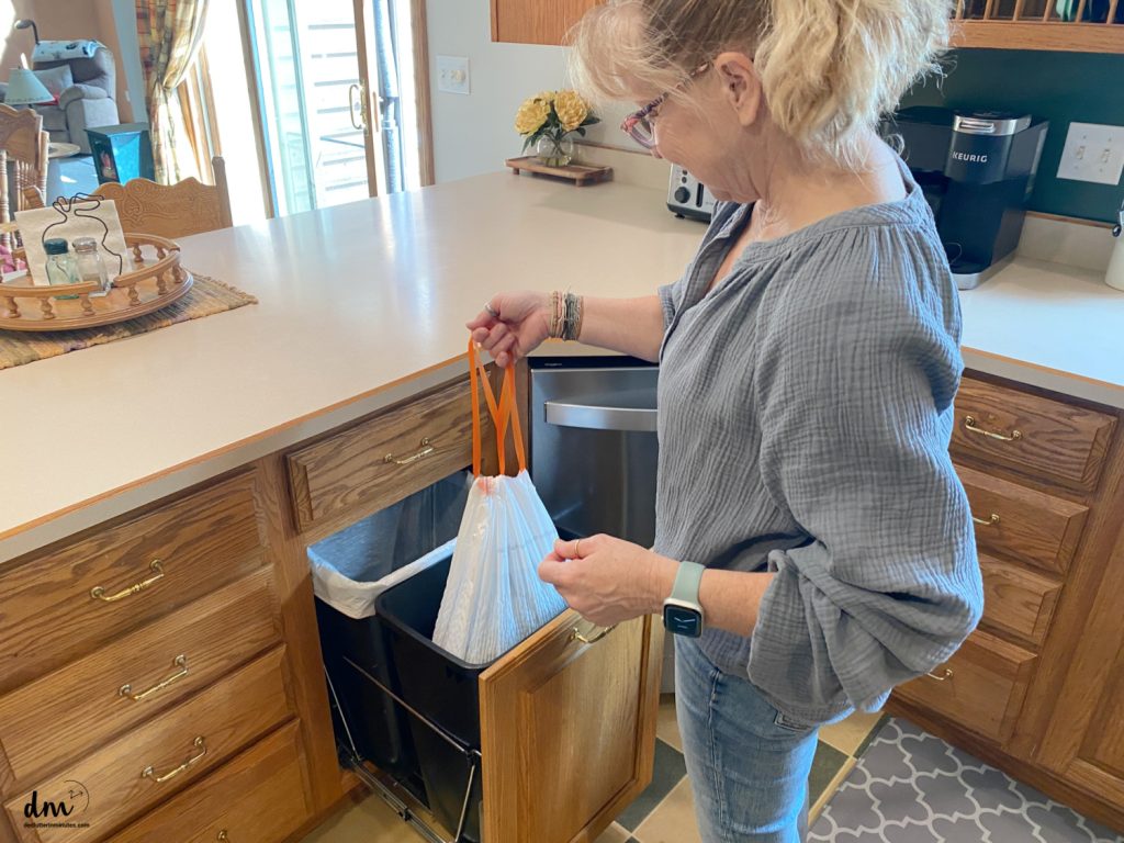 A woman in a kitchen pulling a full trash bag from a built-in cabinet trash bin
