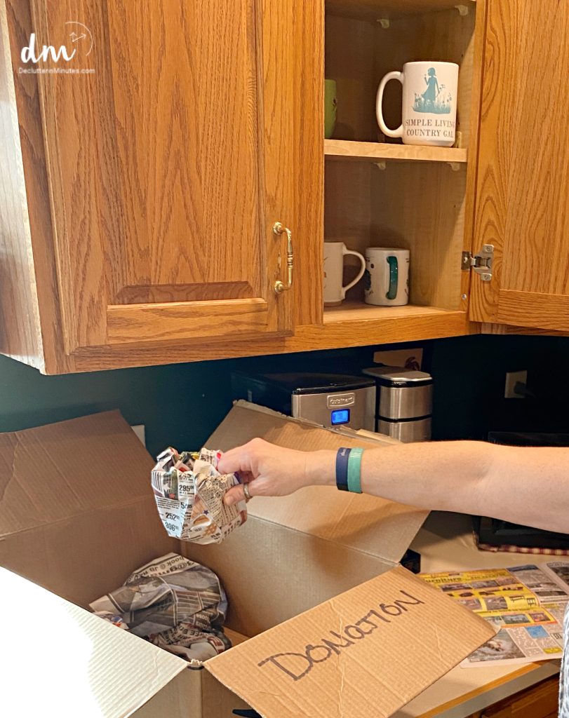 woman reaching into a cabinet decluttering coffee mugs with a donation box