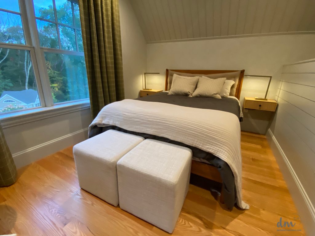 a brown bed with two white foot stools in next to a large open window