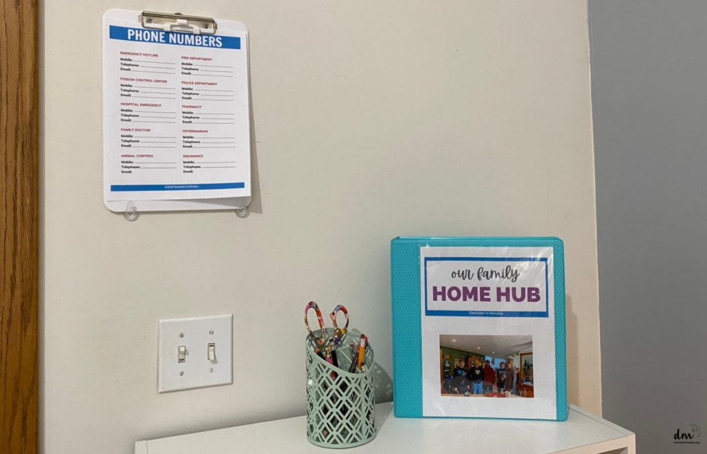 a phone number sheet on a clipboard hanging on a wall next to a binder and pens