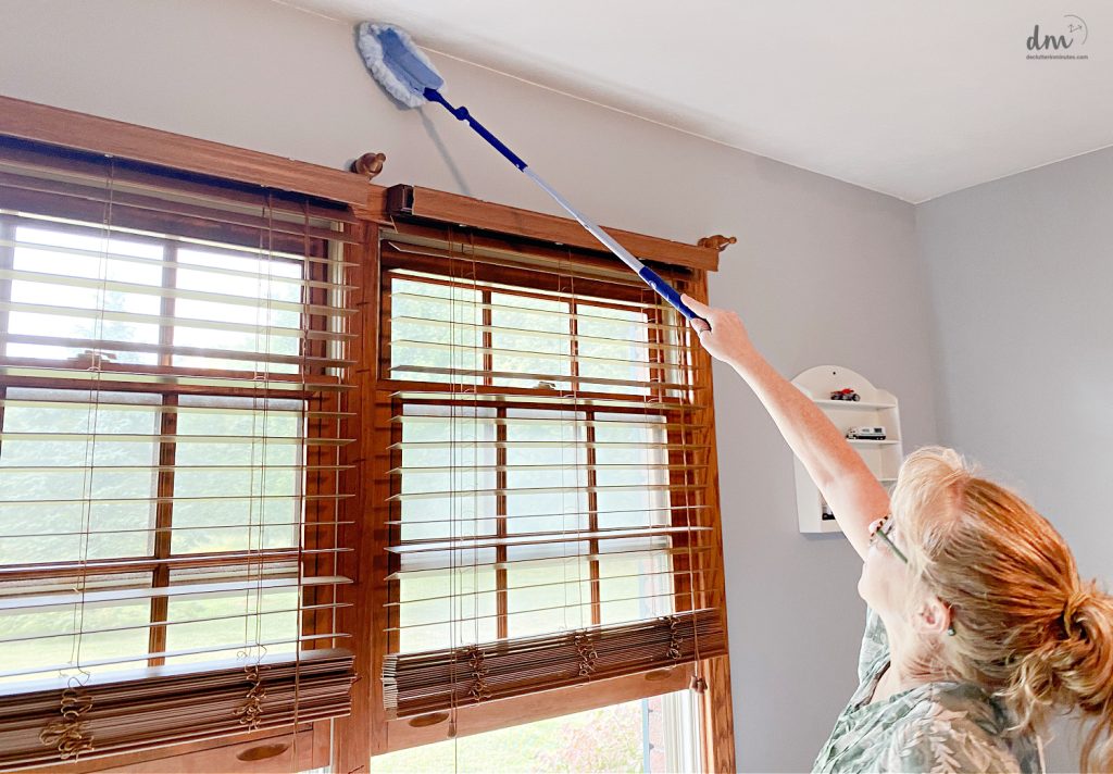 a woman using a duster to clean above a window in a bedroom