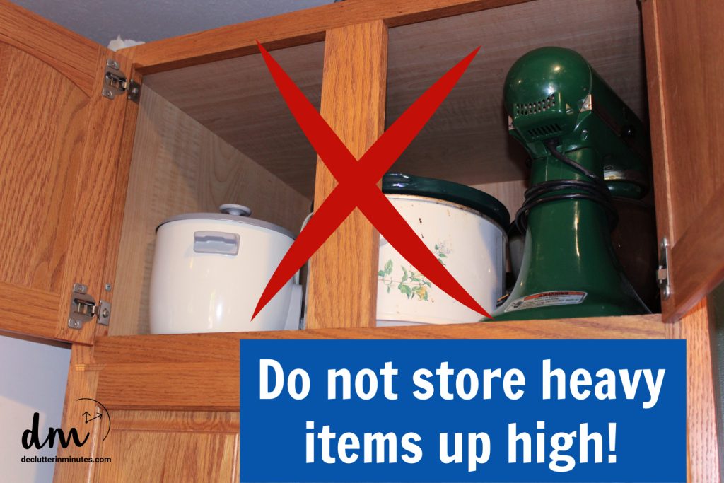 An open wooden kitchen cabinet with a rice cooker, a pot, and a stand mixer with a large red X and a sign that says, "Do not store heavy items up high!"
